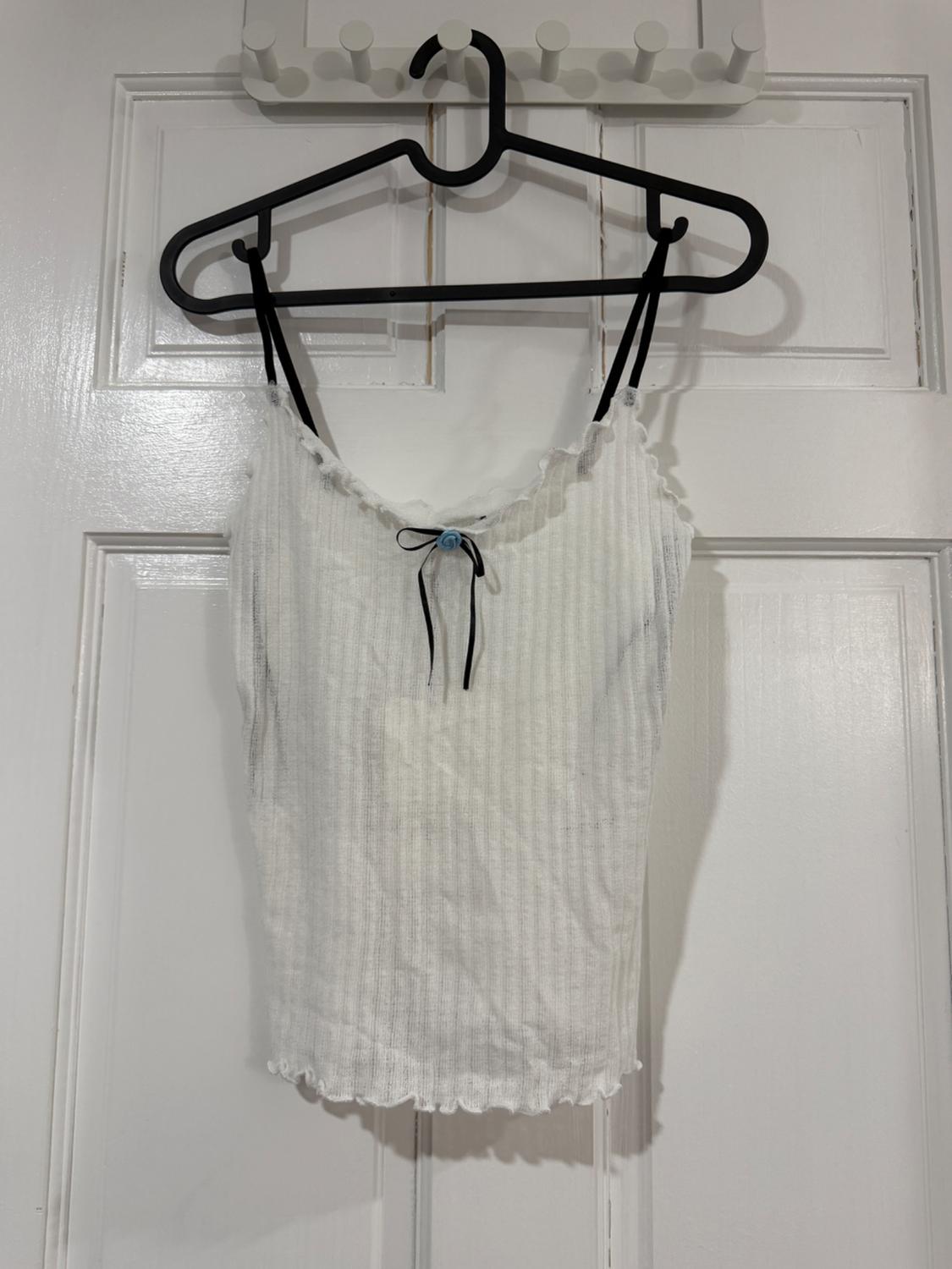 White camisole top with thin straps, a black bow, and a blue detail in the centre, hanging on a black hanger over a white door.