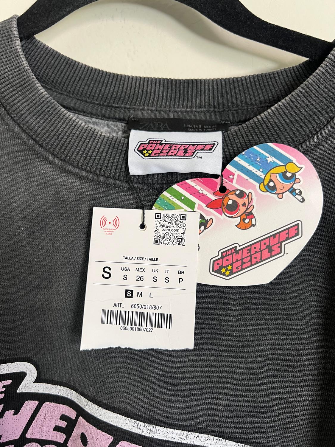 Grey sweatshirt with a "The Powerpuff Girls" tag and sizing. Colourful design with animated characters on the hang tag.