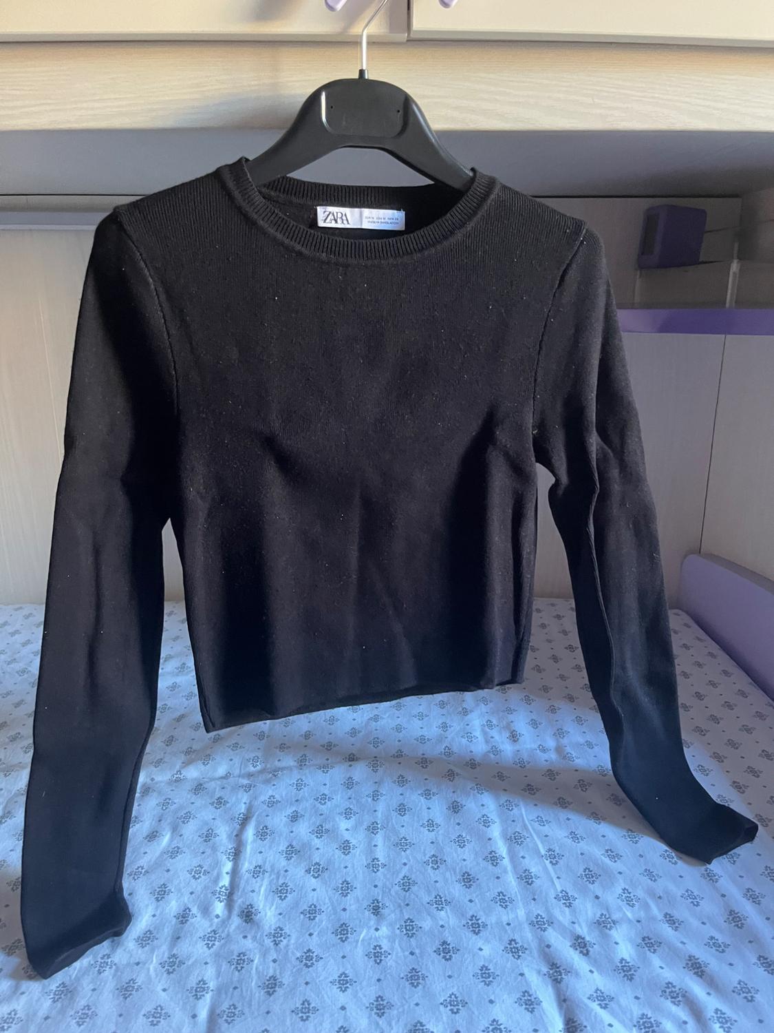 Black long-sleeved, round-neck jumper hanging on a hanger over a bed with patterned white sheets. Text: ZARA.