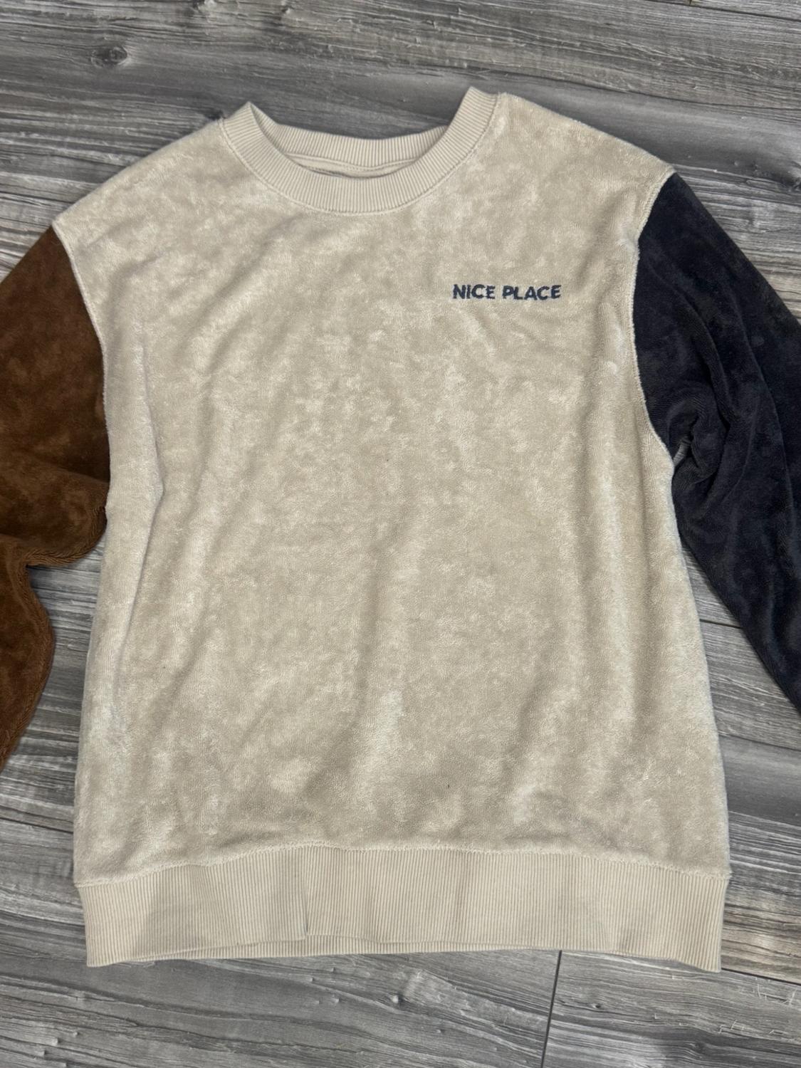 Beige sweatshirt with a soft texture, brown and black sleeves, on a wooden floor. Includes the text NICE PLACE on the chest.