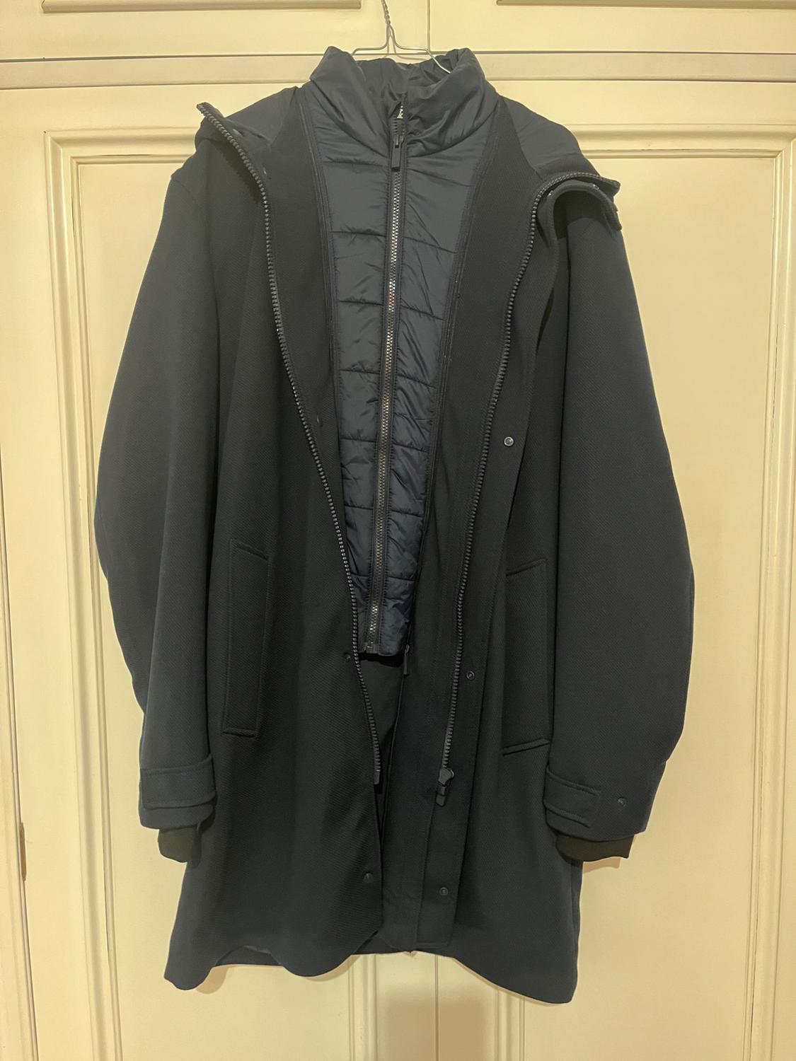Dark blue coat with a zip and hood, hanging on a light wooden door. The inside is padded and also blue.