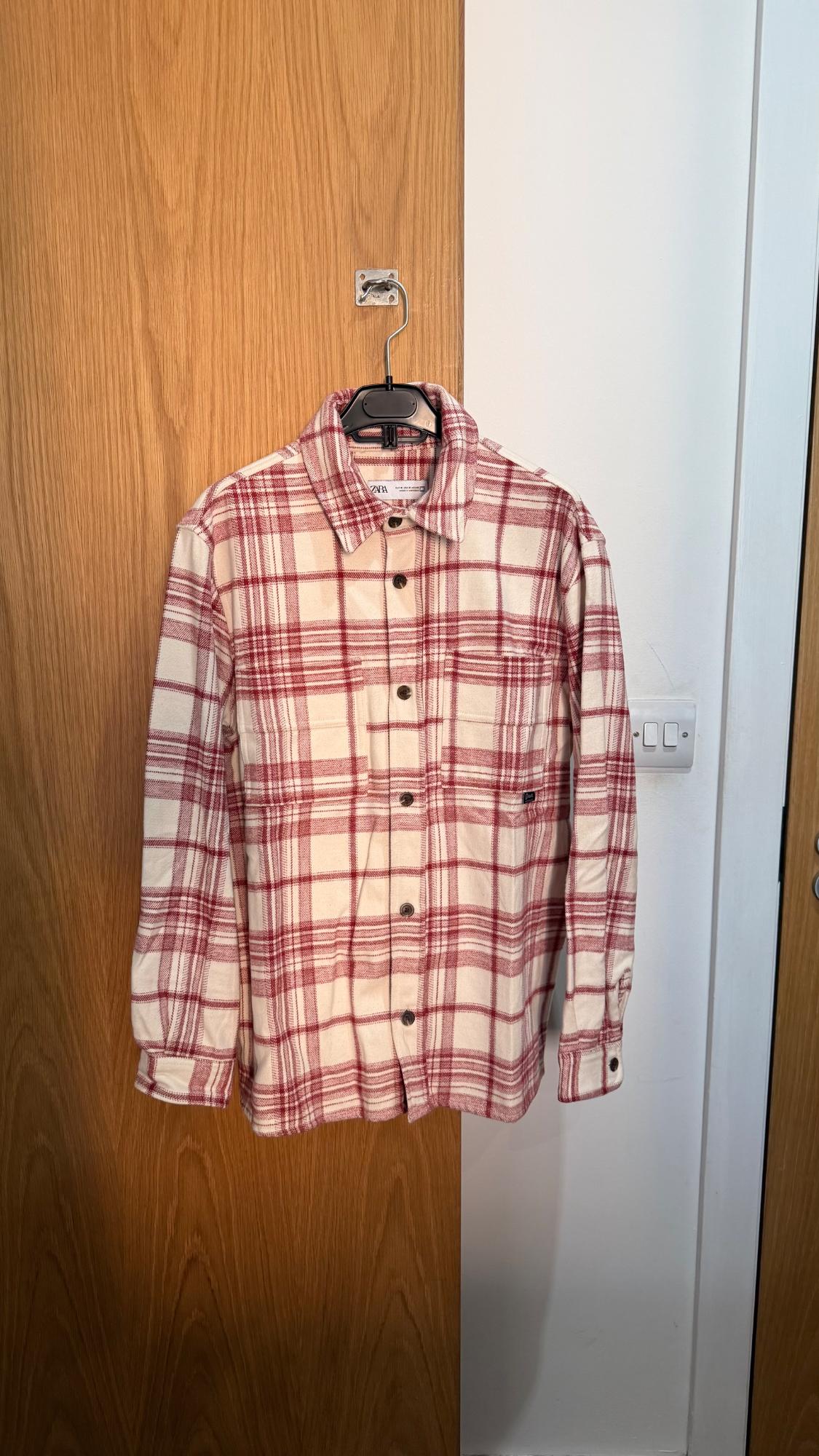 Red and cream checked long-sleeved shirt with a ZARA label, hanging on a wooden door next to a white wall.