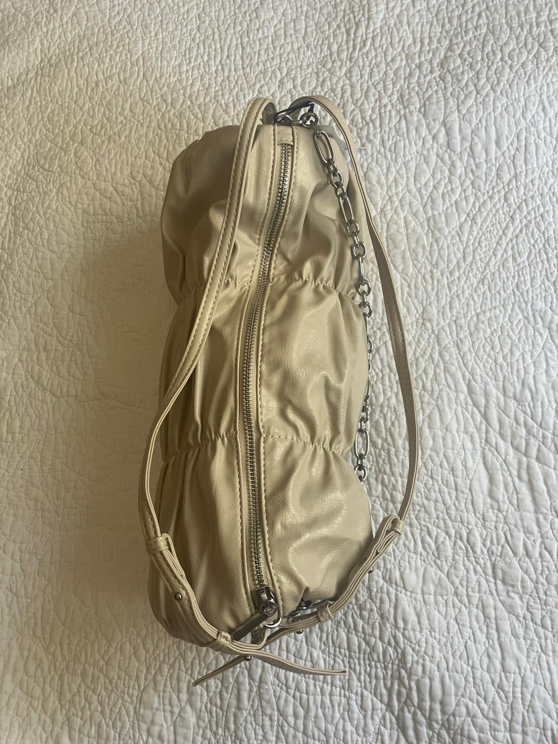 Beige bag with a crinkled texture and central zip, on a white bedspread. Metal chain handle and leather strap.