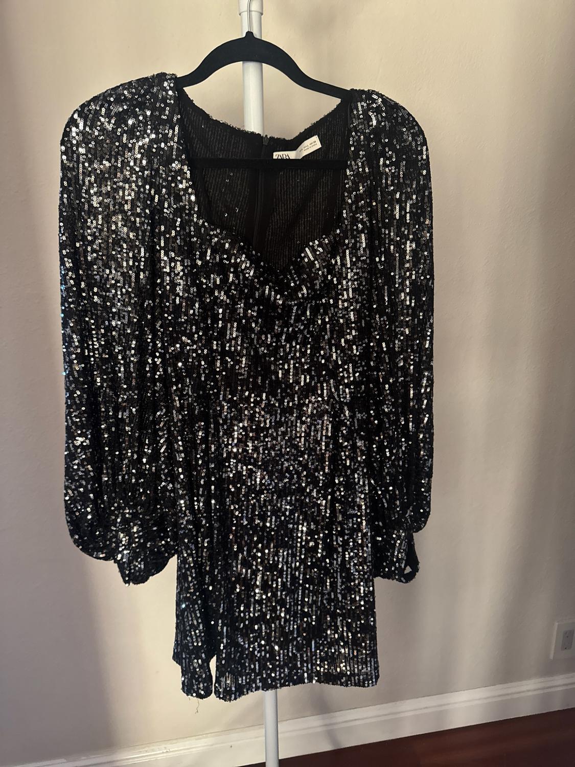 Short black sequined dress with long puff sleeves hanging on a black hanger. Light-colored wall background. Tag with text ZARA.