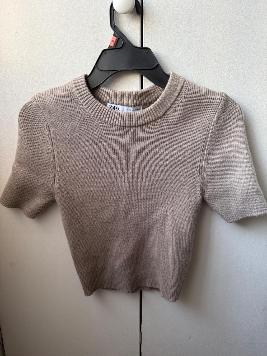 Beige, short-sleeved, crew neck knit top, hanging on a black hanger in front of a white closet. Tag with ZARA text.