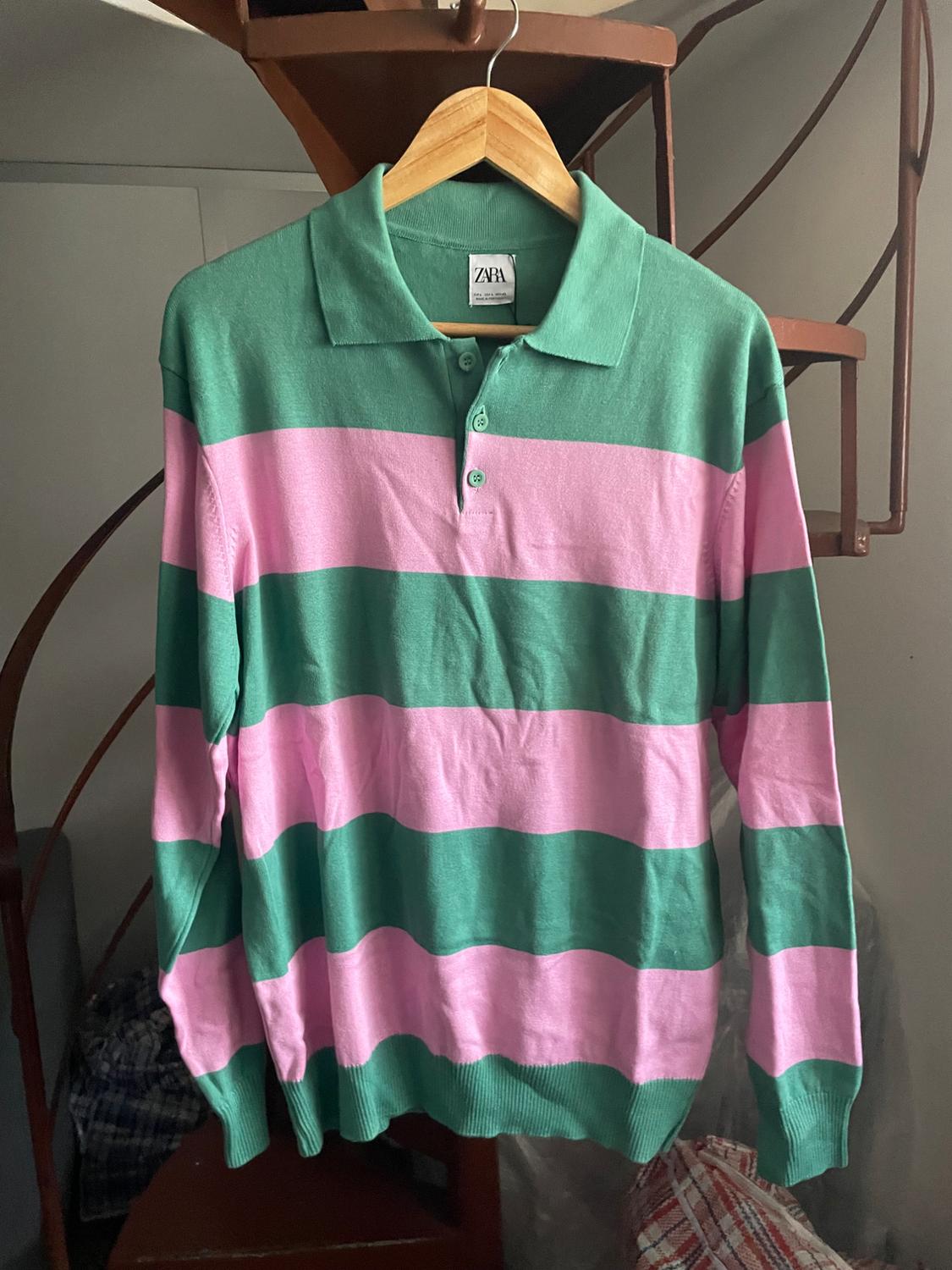 Green and pink striped jumper hanging on a wooden hanger. Background with spiral staircase and bags. Visible label: Zara.