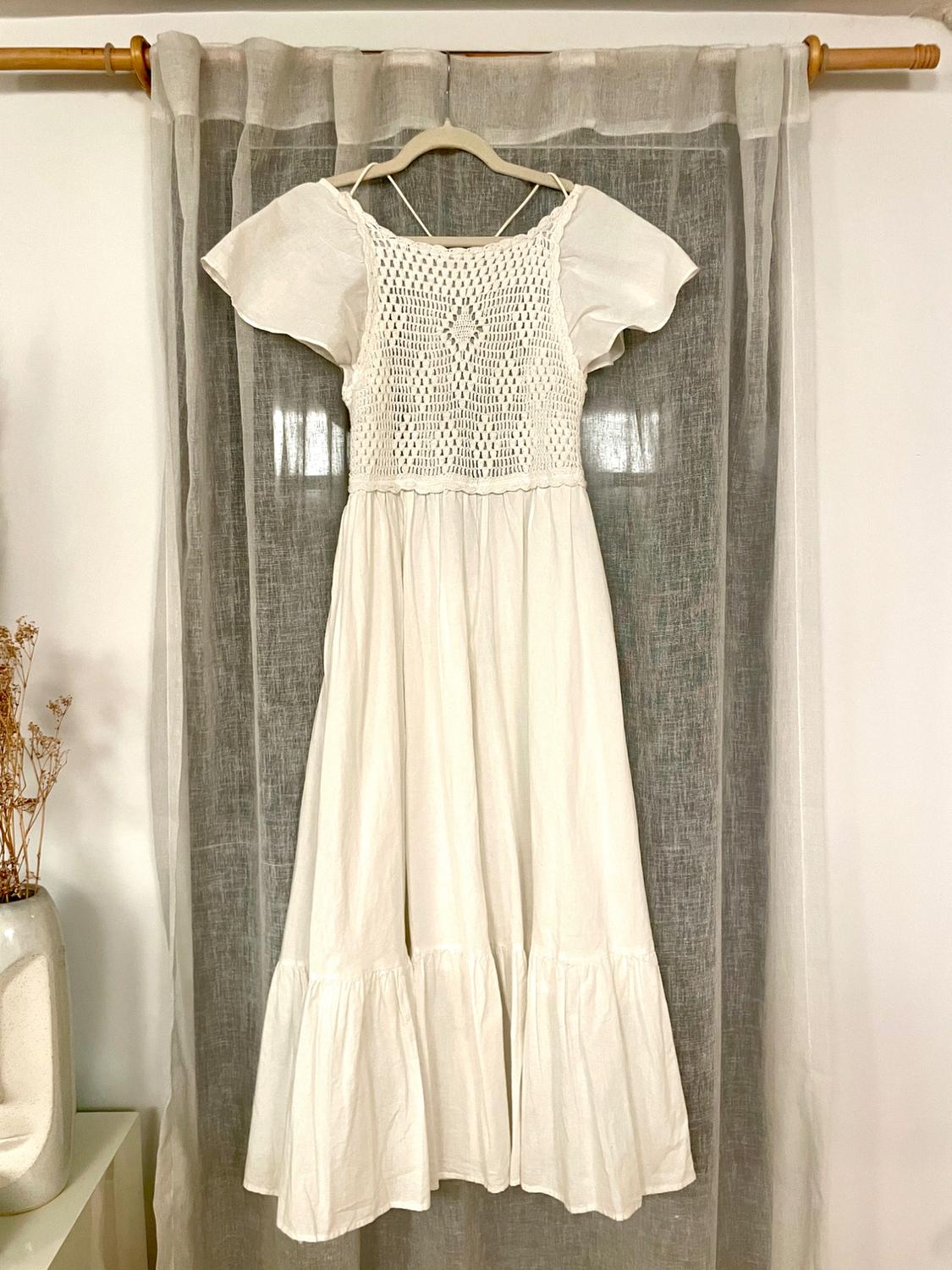 White lace dress hanging on a hanger in front of a sheer curtain. To the left, a vase with dried flowers.