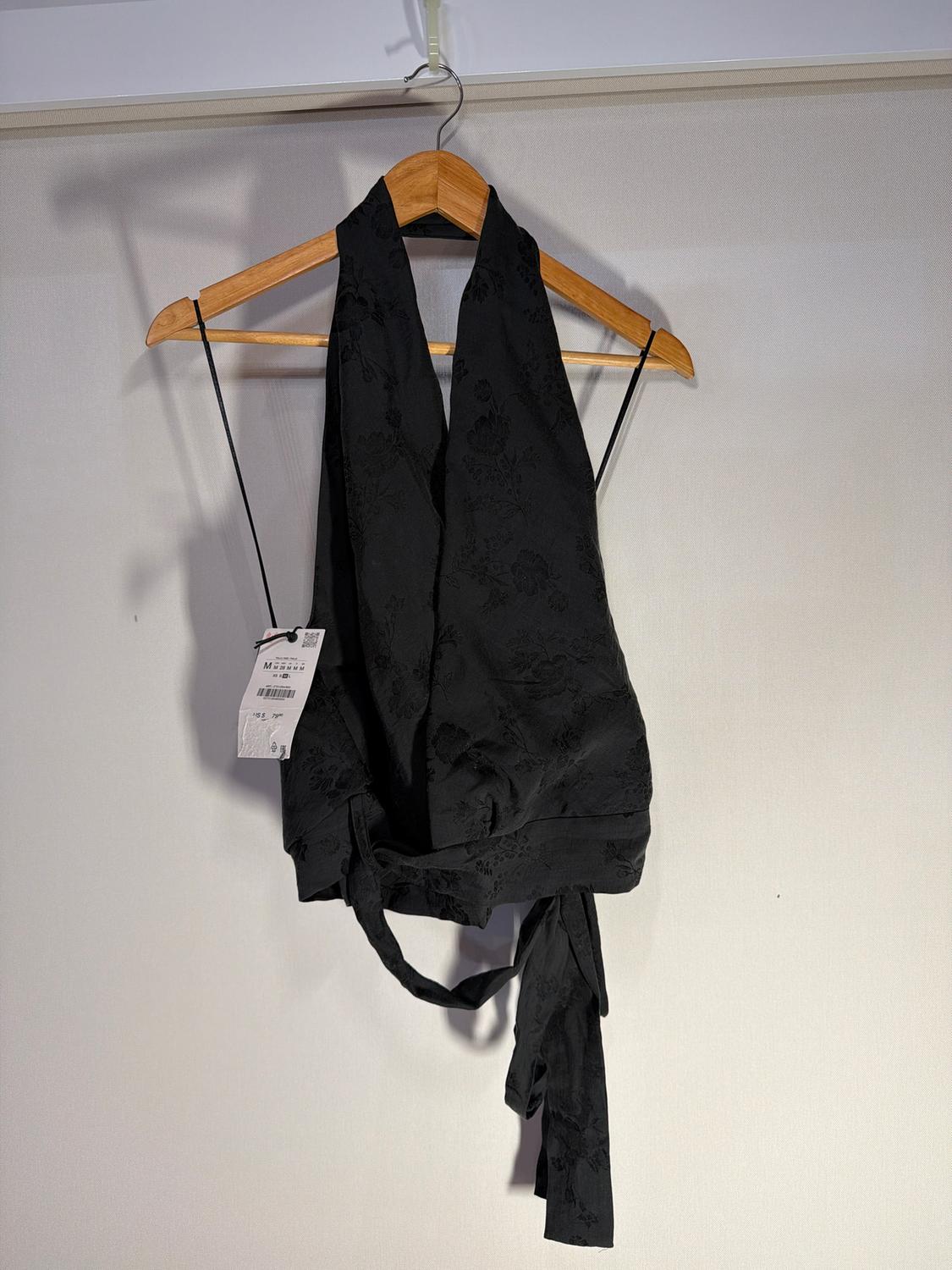 Black floral print top hanging on a wooden hanger. White label visible with product details. Light background.
