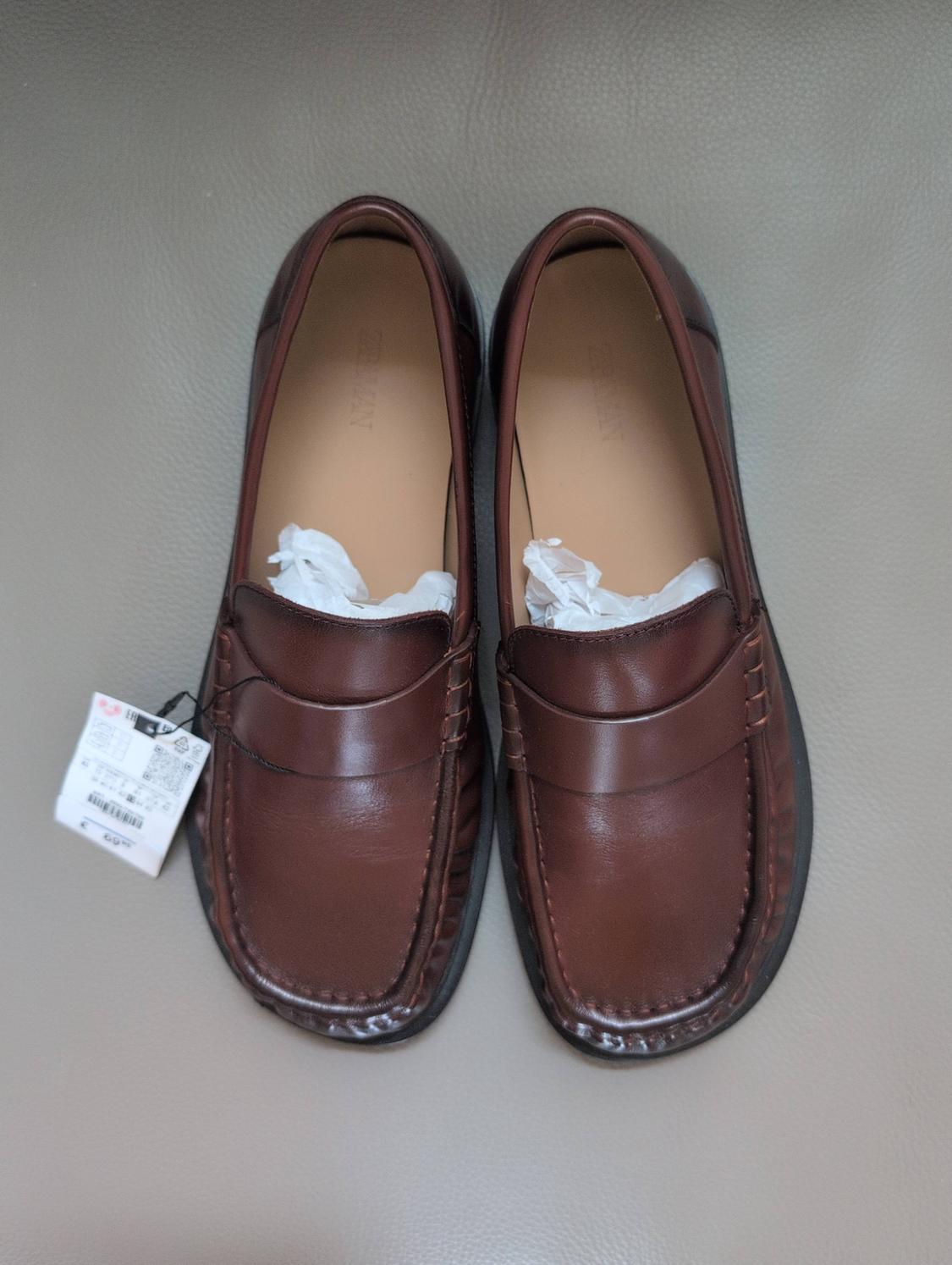 Top view of a pair of brown loafers on a grey background. They have paper inside and a label. Text: ZARA MAN.