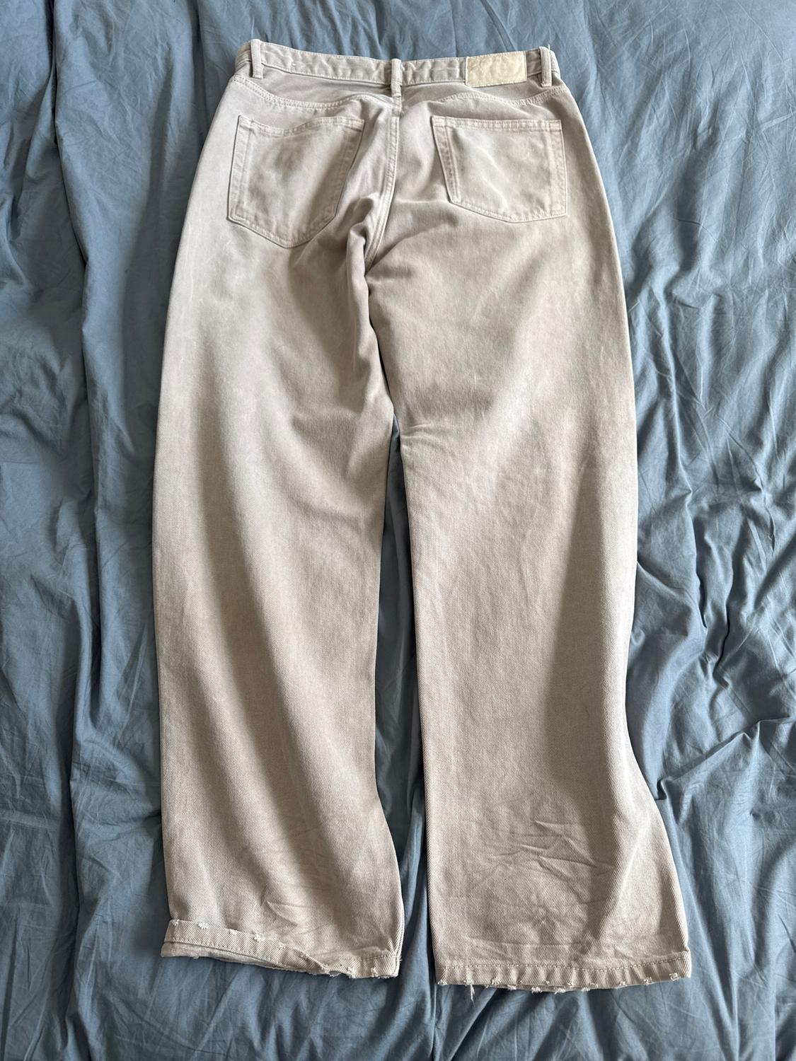 Straight-cut beige trousers with back pockets, placed on a bed with wrinkled blue sheets.