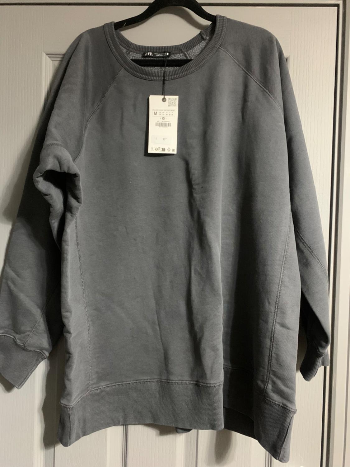 Grey long-sleeved sweatshirt hanging on a hanger, with visible label indicating size M, against a white door background.