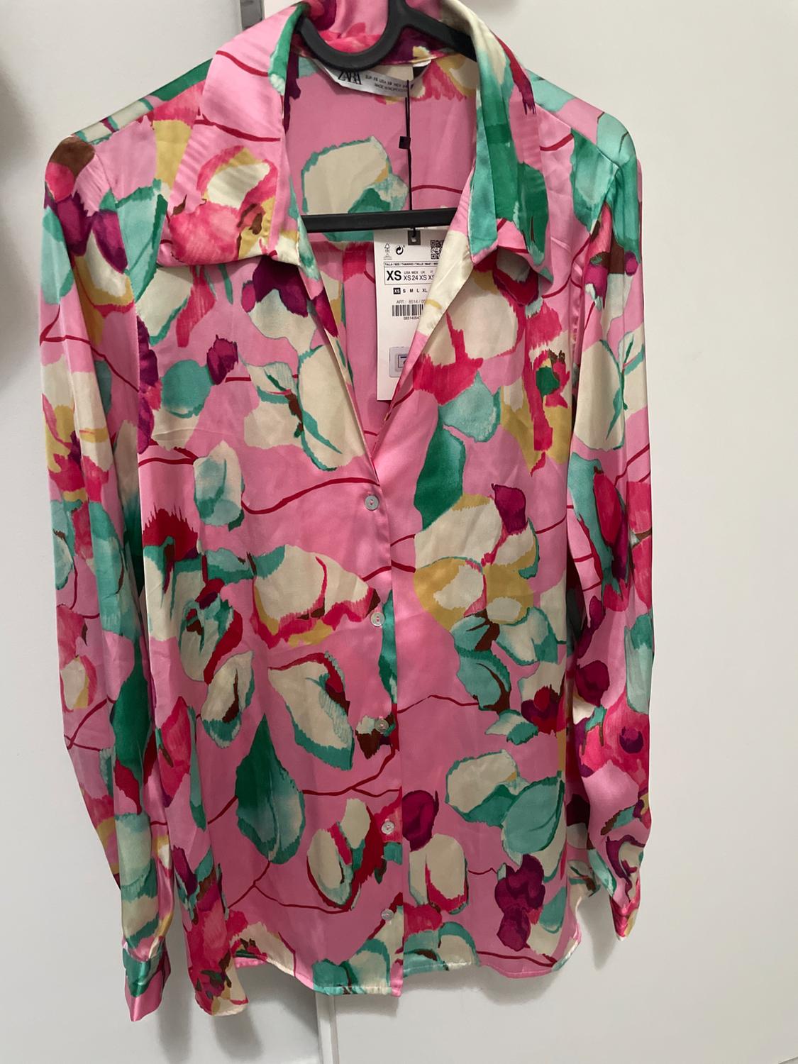 Long-sleeved shirt with a floral print in pink, green, and cream tones, hanging on a hanger with a visible XS size tag.