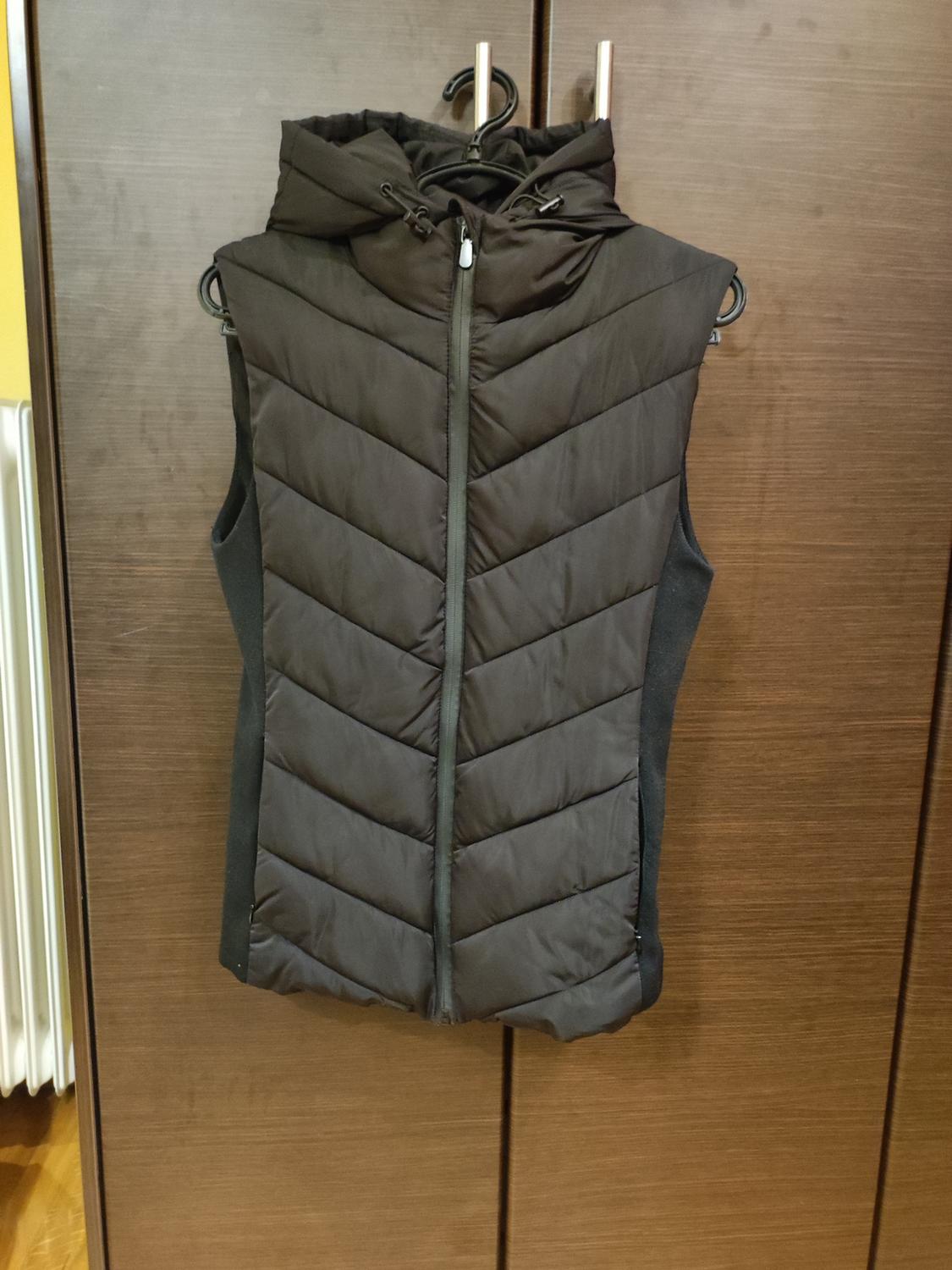 Black padded gilet with hood and zip, hanging on a hanger in front of a wooden wardrobe.