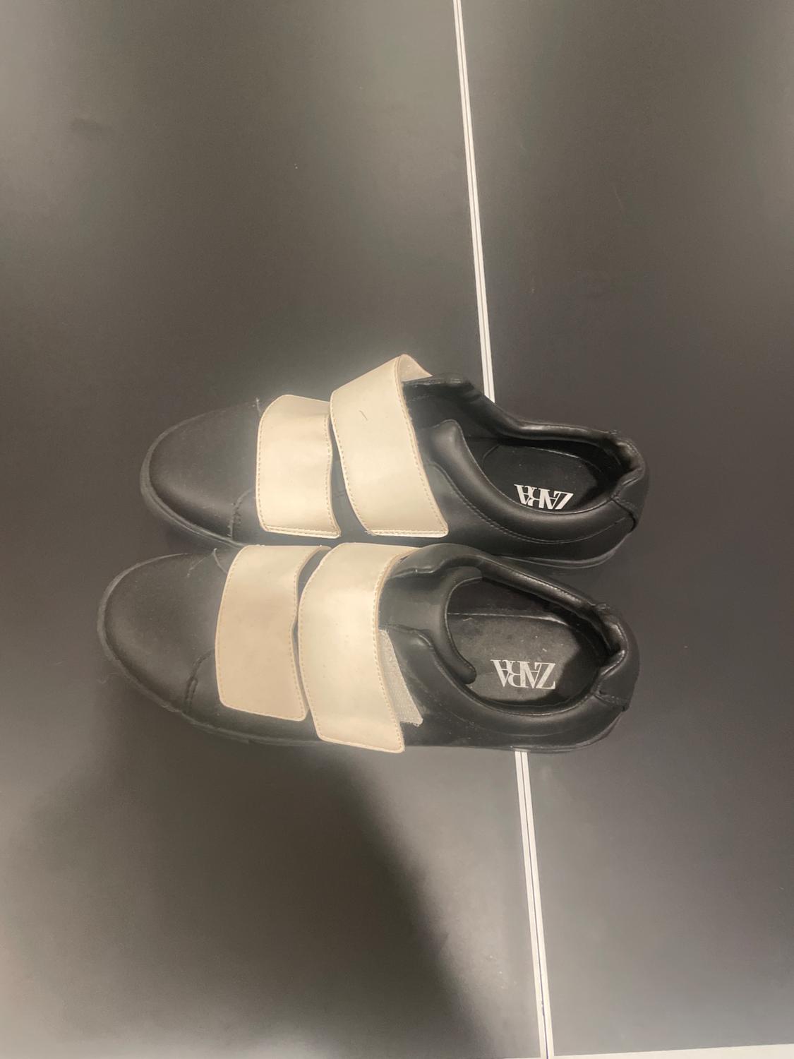 Black sneakers with two white stripes on a dark surface with a white line. Inside, the text ZARA can be read.