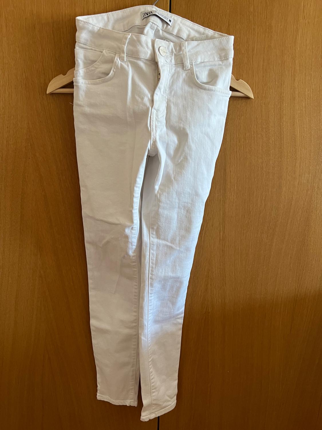 White jeans with ZARA tag, hanging on a wooden hanger against a wooden background.