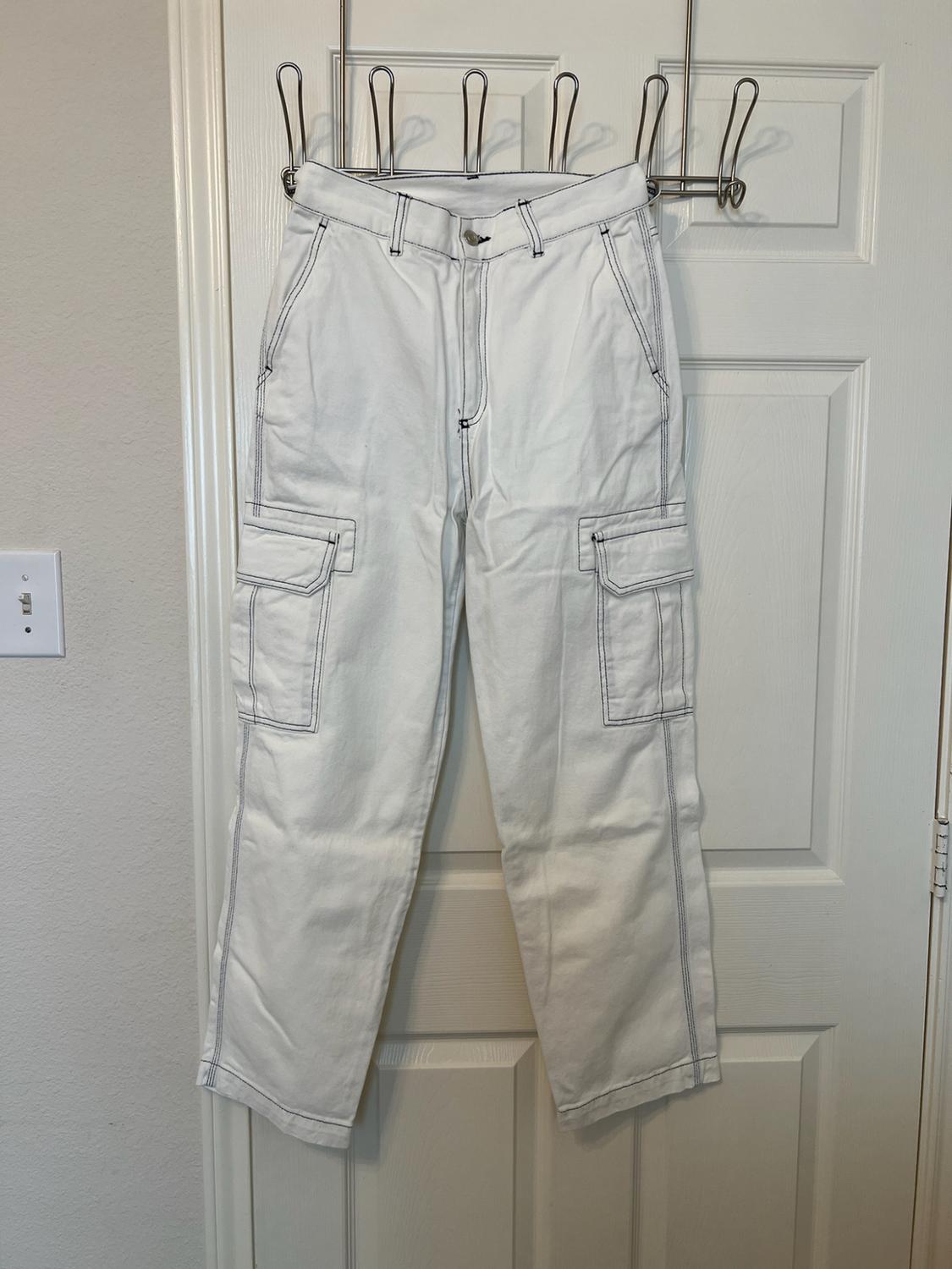 White cargo trousers with contrasting dark stitching, hanging on a metal hanger over a white door.