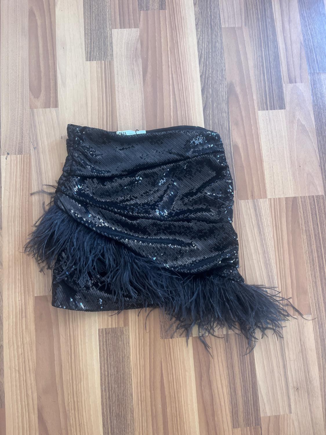 Black sequin skirt with feathers on the bottom hem, on a light wooden floor.