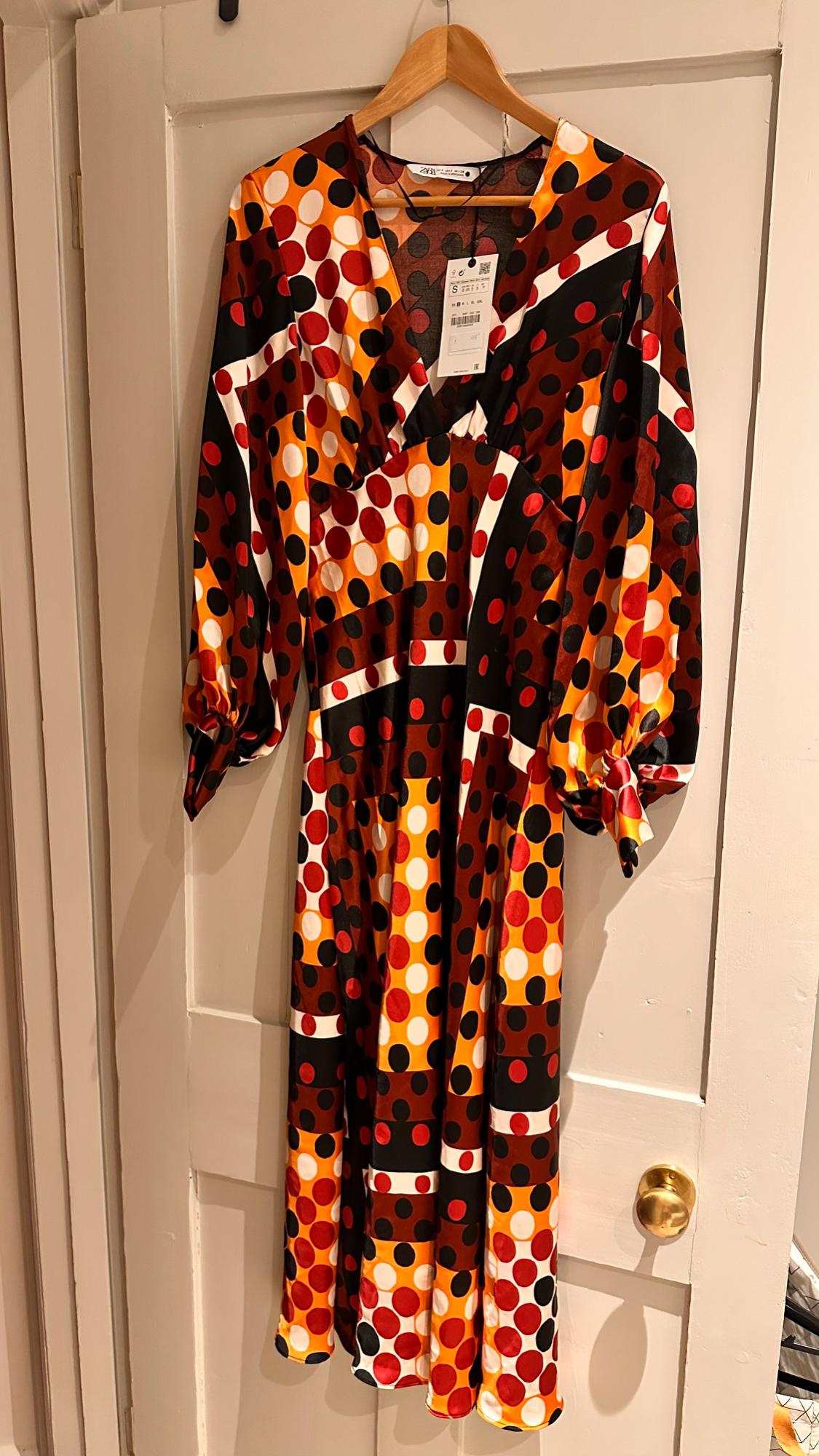 Dress hanging with a polka dot print in red, orange, black and white. Label visible on the neck. White door background.
