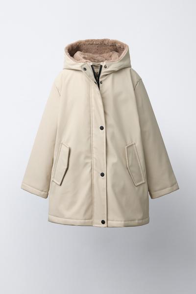 Frontansicht einer sandfarbenen Jacke mit Kapuze, Reißverschluss- und Knopfleiste, Pattentaschen und Innenfutter in Felloptik.