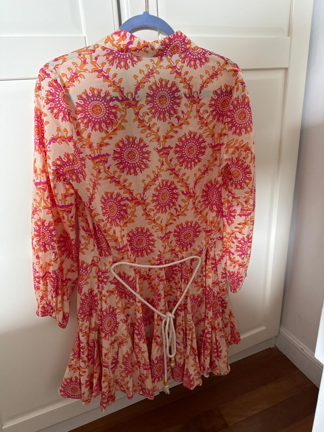 Short dress with a pink and orange print, long sleeves and a white rope belt, hanging on a hanger in front of a white wardrobe.