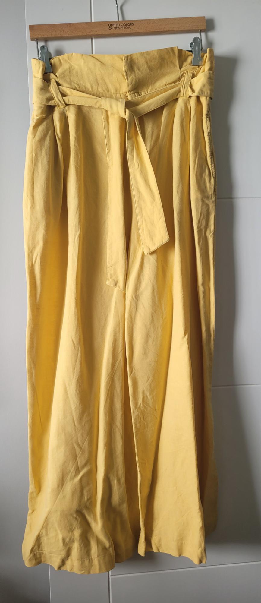 Yellow, light fabric trousers with a belt, hanging on a wooden hanger that says "United Colors of Benetton".