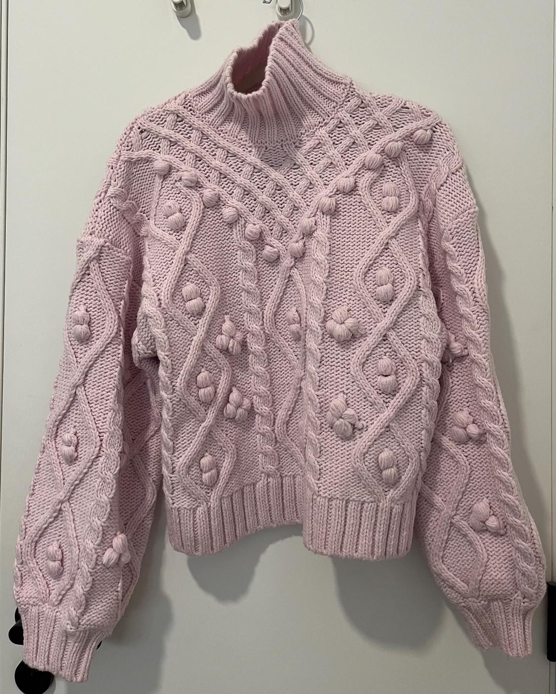 Chunky pink high-neck knitted jumper with cable knit and pom-pom design, hanging on a white hanger against a light background.