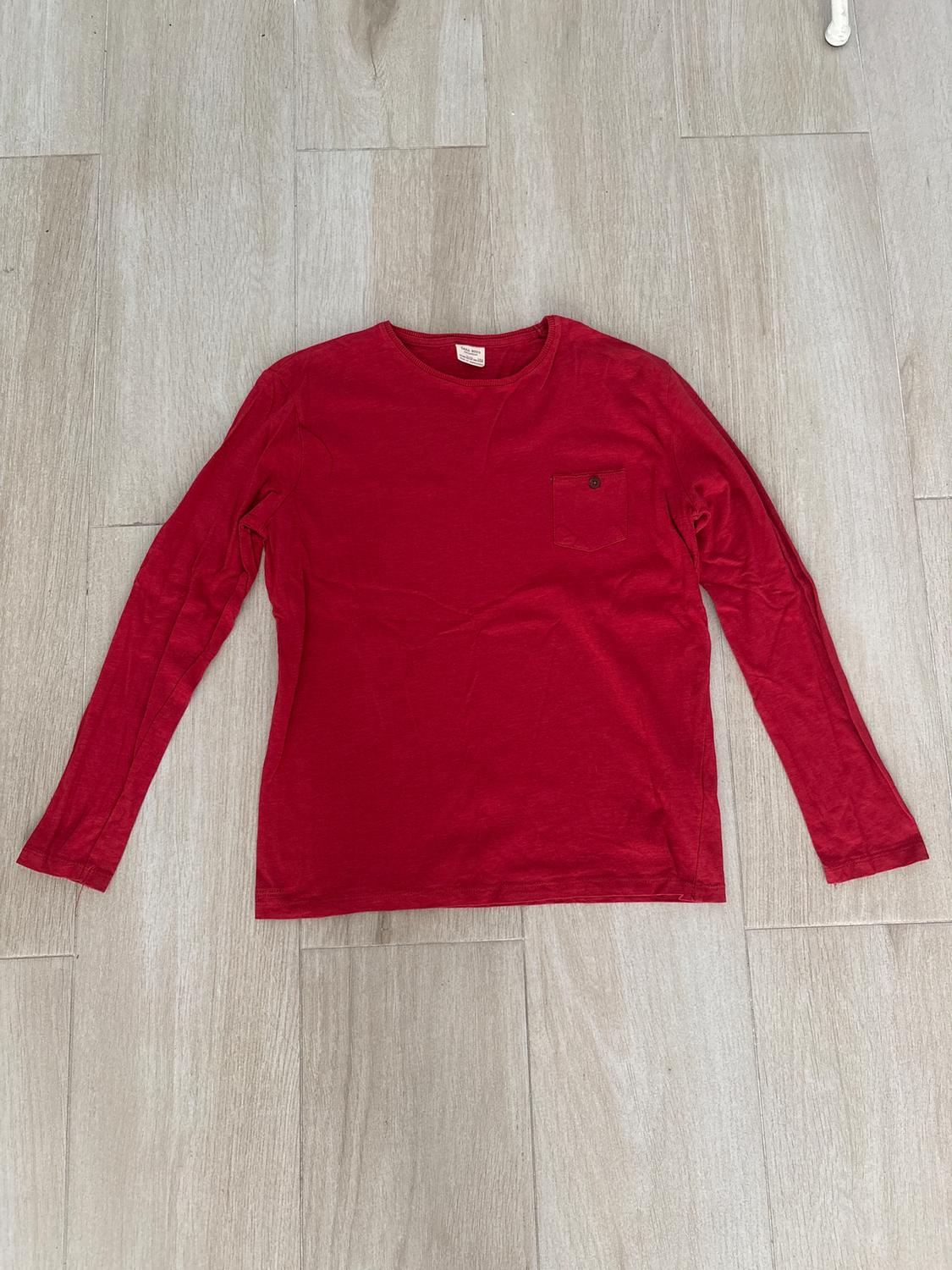 Red long-sleeved T-shirt with chest pocket, on a light wooden floor. Visible label on the collar.