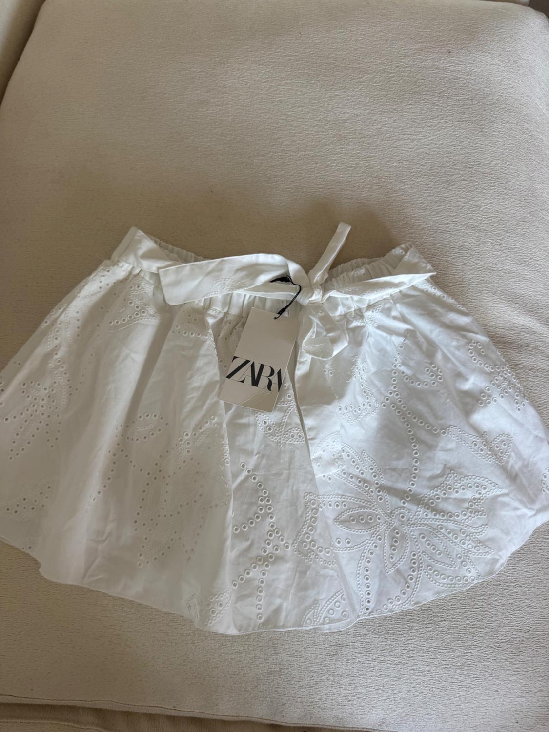 White skirt with floral embroidery and ZARA brand tag, on a beige background.