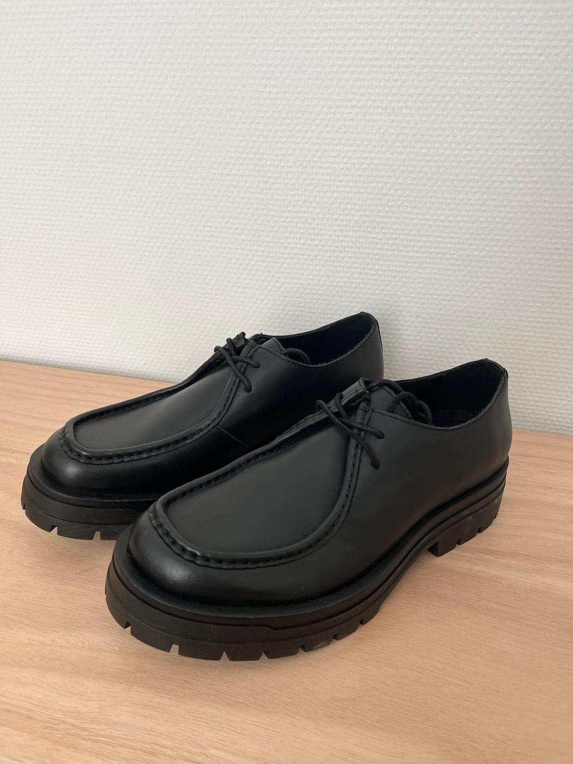 Pair of black lace-up shoes with a thick sole, resting on a light wood surface in front of a white wall.