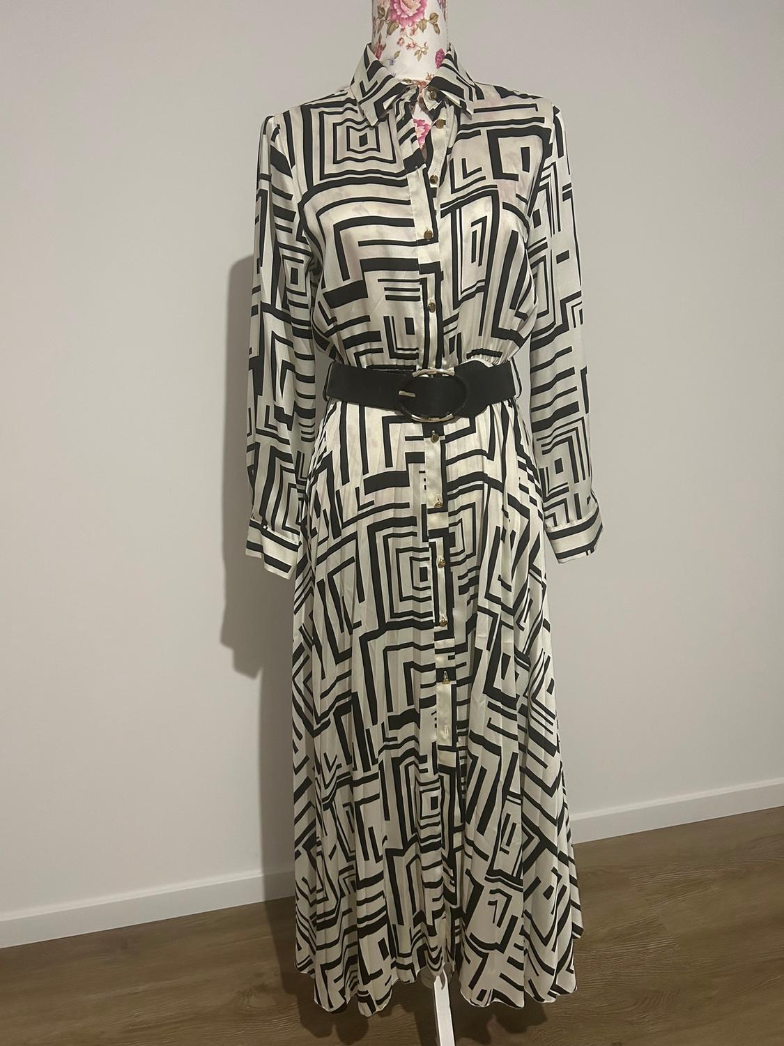 Long dress with geometric black and white print, black belt, displayed on a mannequin with a white background and wooden floor.