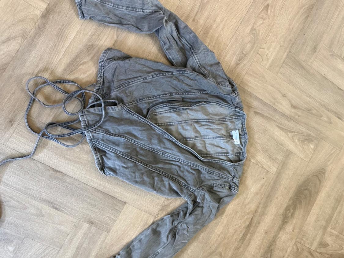 Grey long-sleeved denim top with visible stitching and tie details, laid out on a herringbone wooden floor. Casual and relaxed style.