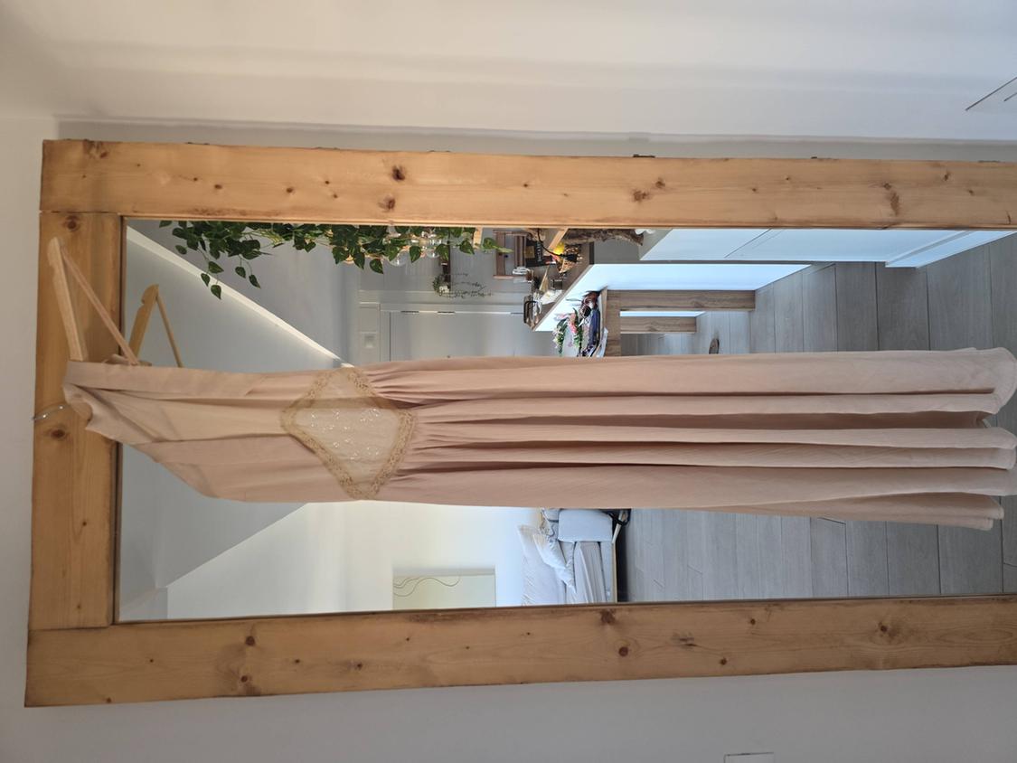 Beige maxi dress with lace detailing hanging in front of a rustic wooden mirror reflecting a bright room.
