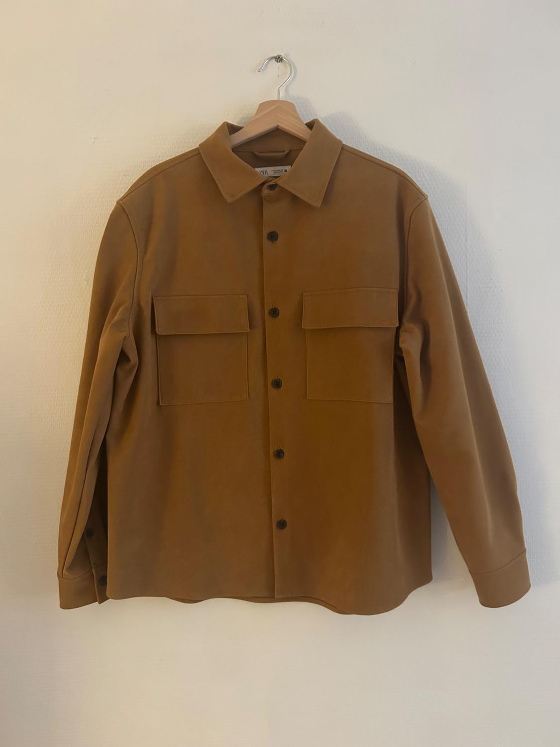 Brown long-sleeved shirt with black buttons and two front pockets, hanging on a hanger against a white background.
