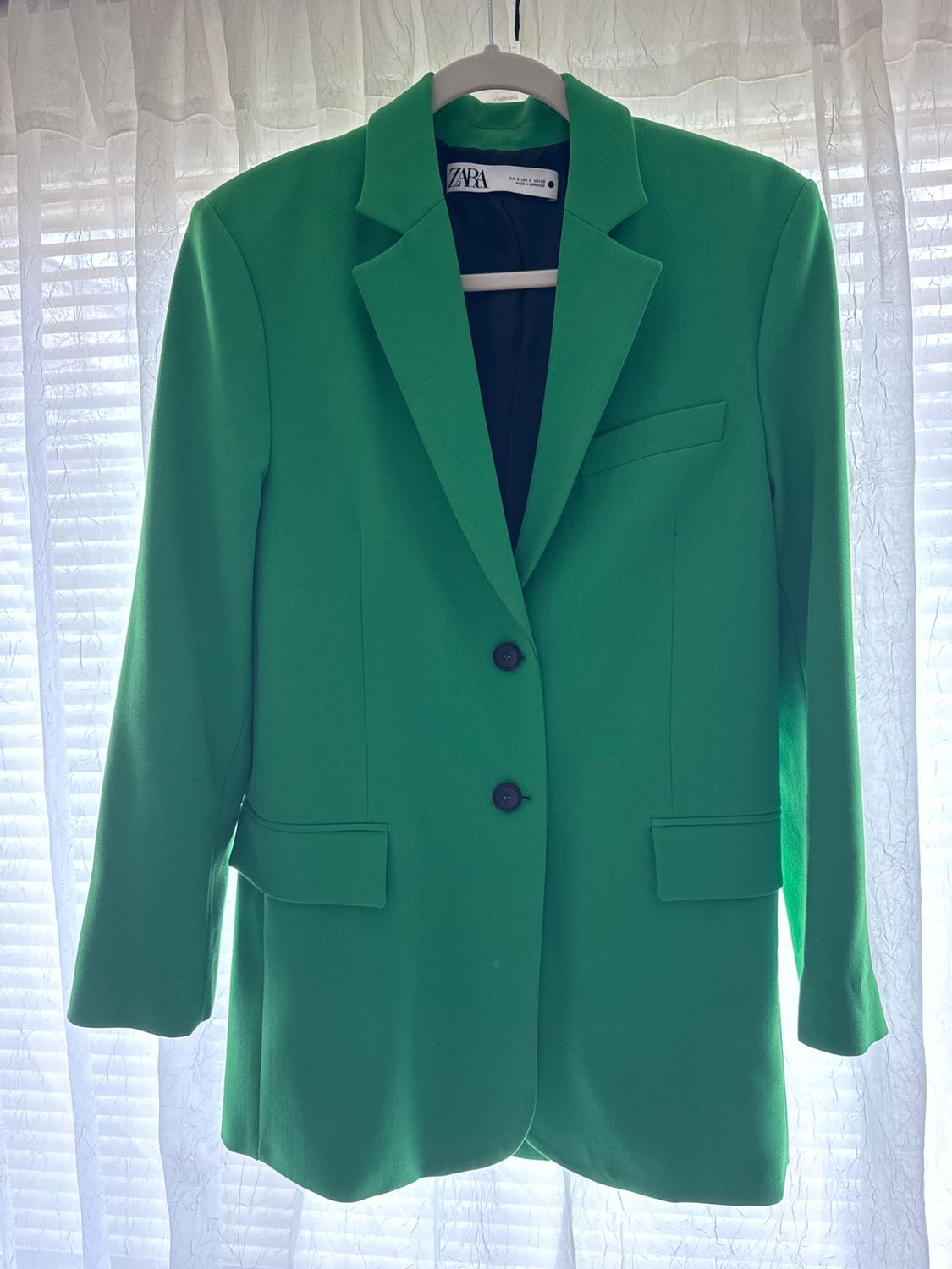 Green jacket hanging on a hanger in front of a white curtain. Visible tag with the text "ZARA".