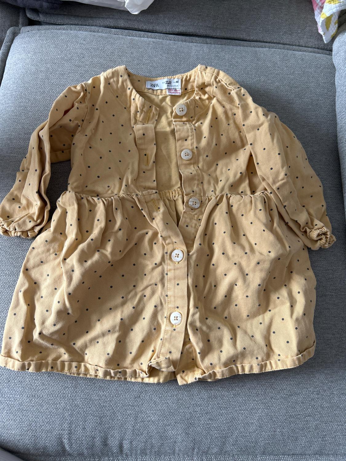 Child's beige dress with black polka dots and white buttons, on a grey sofa. Label visible on the collar.