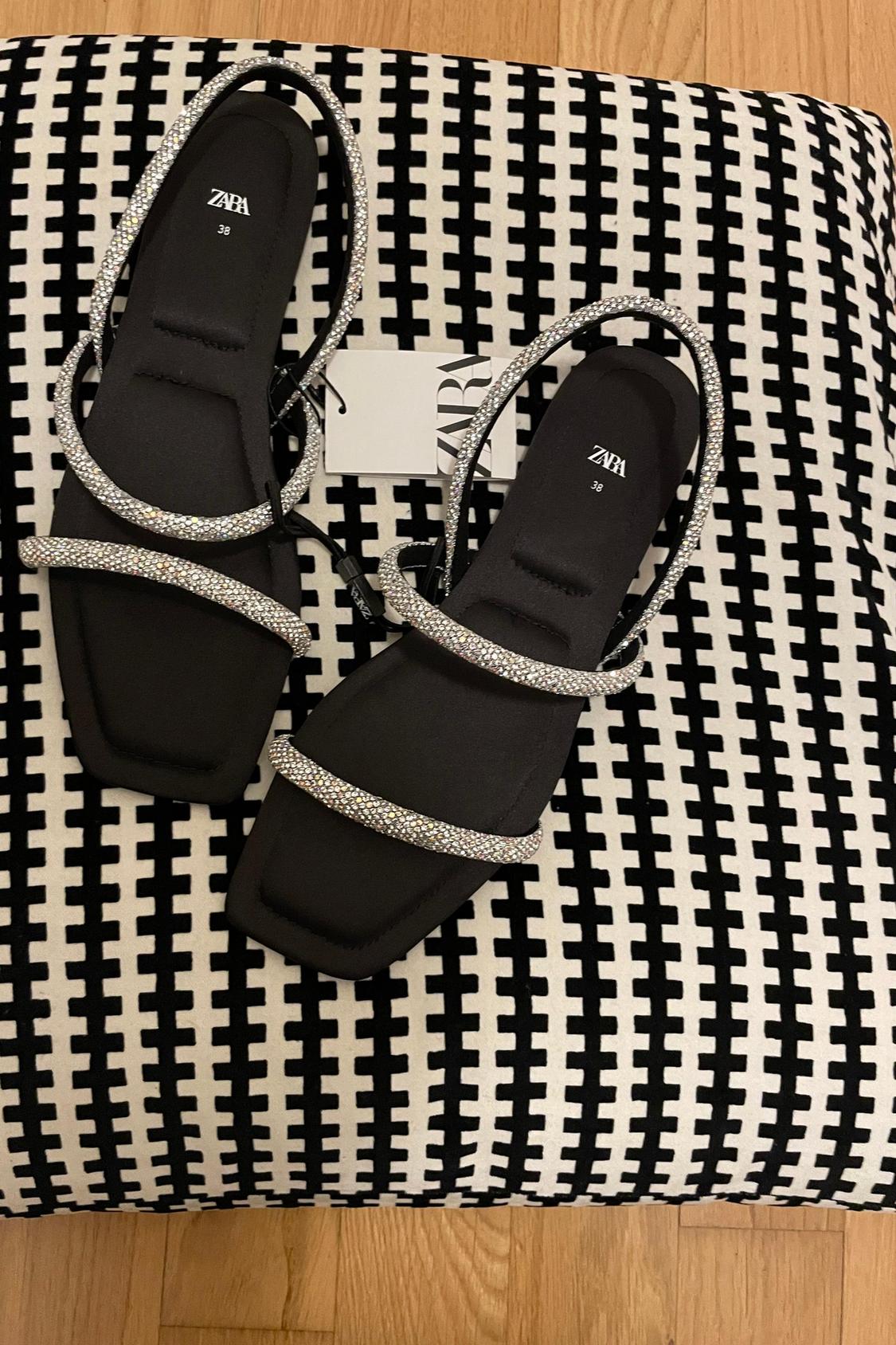 Black sandals with sparkling straps and visible label, on a black and white patterned background.
