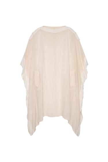Cream-colored loose tunic with white trim, two front pockets, and an asymmetrical hem, presented against a white background.