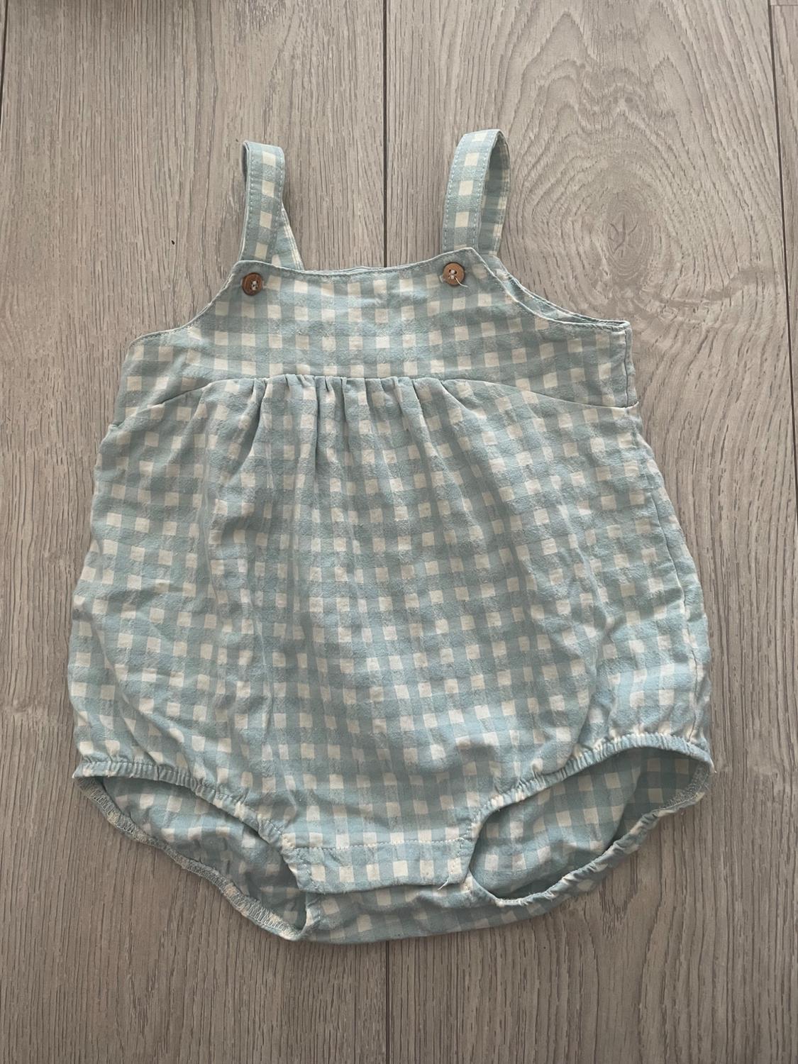 Blue and white checked baby romper with straps and wooden buttons, on light wooden floor.