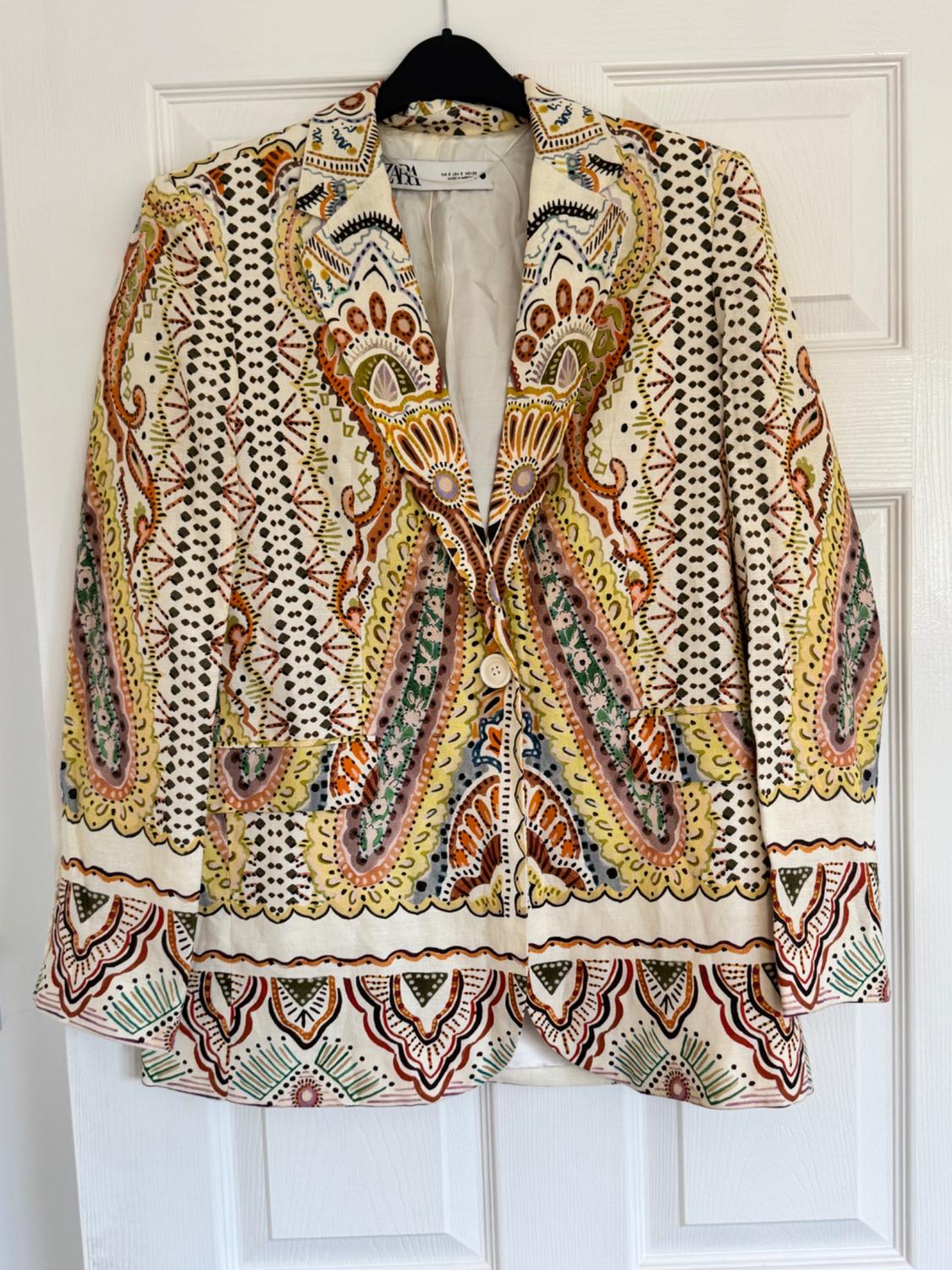 Printed jacket with geometric and floral patterns in shades of yellow, pink, green and black, hanging on a hanger in front of a white door.