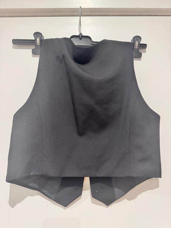 Black waistcoat hung on a hanger against a white background. Sleeveless and round neck design, with an asymmetrical cut on the hem.
