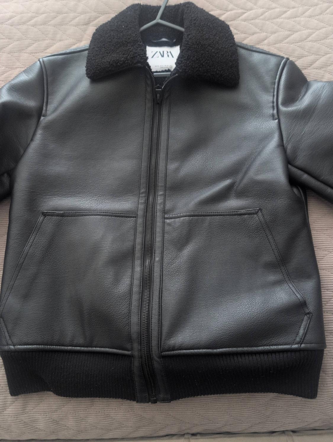 Black jacket with zip, pockets, elasticated hem and faux fur collar. Label with Zara text. Placed on a beige surface.