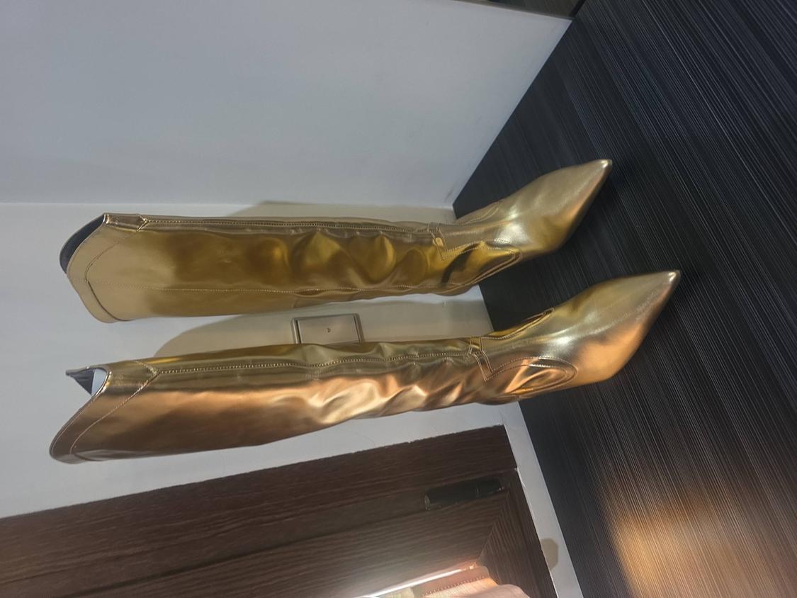 Gold metallic pointed high-heeled boots, arranged horizontally against a white wall and dark panelled background.