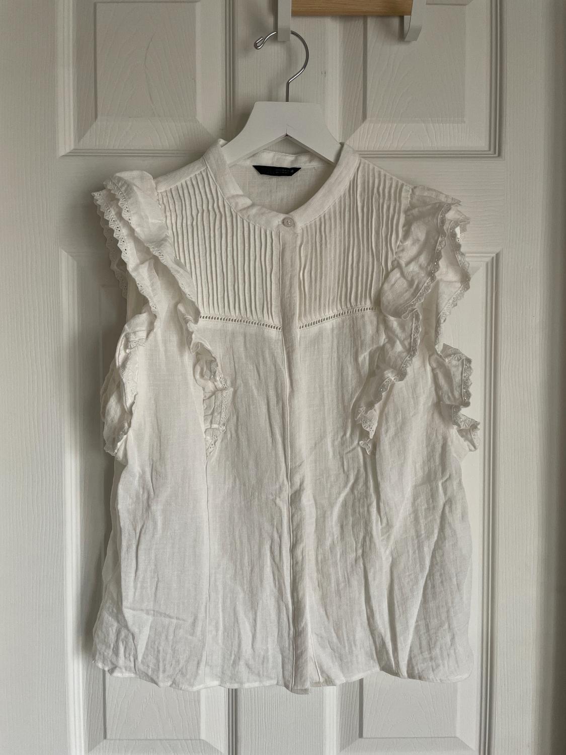 White sleeveless blouse with ruffles and lace details, hanging on a white hanger over a white door.