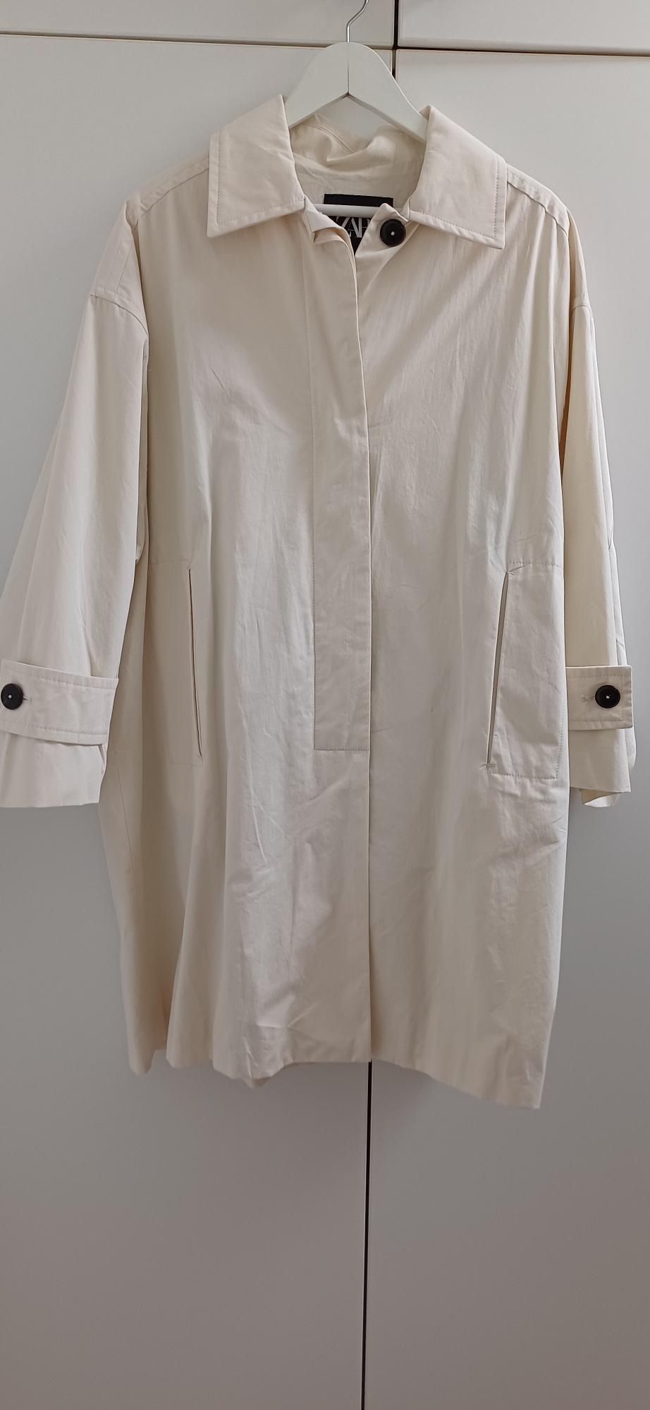 Beige coat hanging on a white hanger, with dark buttons on the cuffs and a classic collar, against a light background.