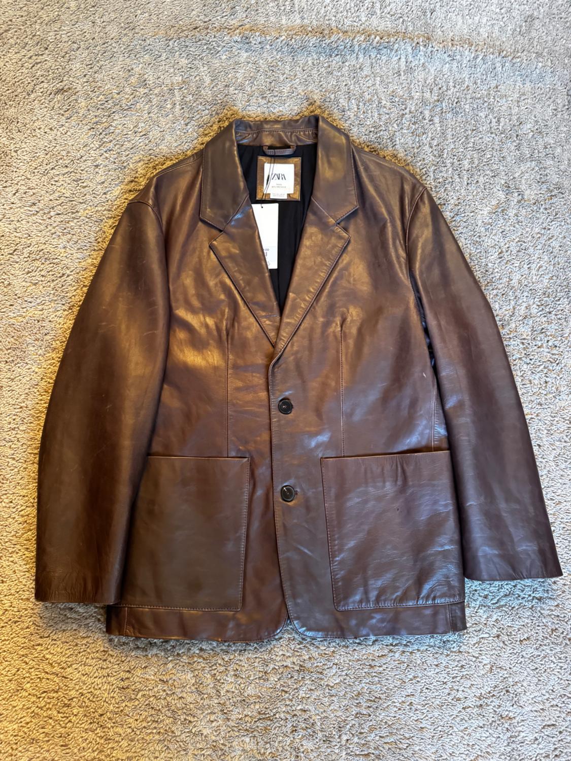 Brown leather jacket with lapels and two buttons, on a beige rug. Visible label on the collar with text "ZARA".
