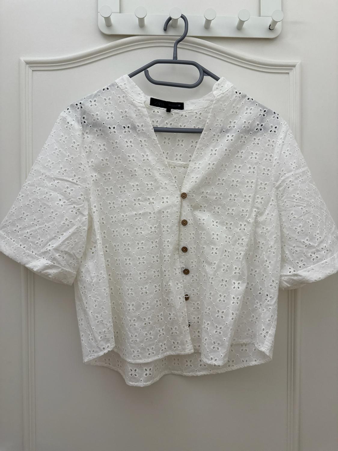 White short-sleeved blouse with cutwork embroidery, V-neck and brown buttons. ZARA label. Hanging on a hanger on a white door.