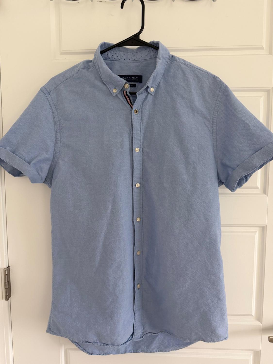 Light blue, short-sleeved button-down shirt, hanging on a black hanger over a white door. The tag reads ZARA MAN.