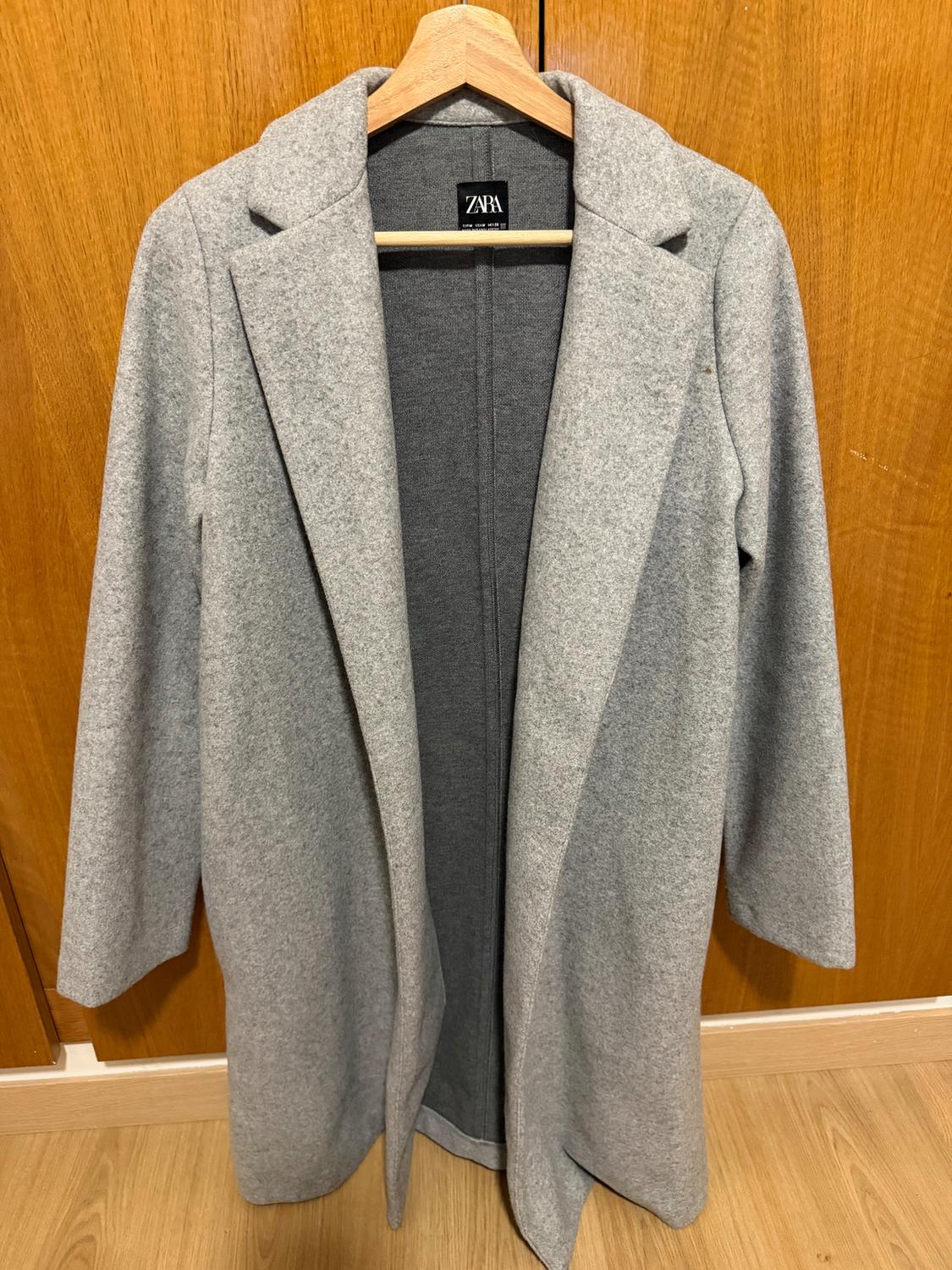 Grey coat hanging on a wooden hanger, with a visible ZARA brand label, against a background of wooden doors.