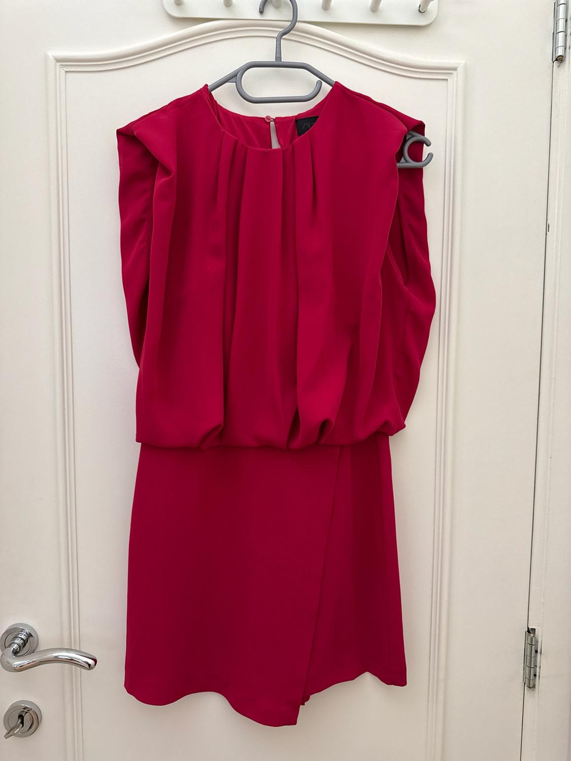 Fuchsia sleeveless short dress with a draped bodice and wrap skirt, hanging on a hanger over a white door.