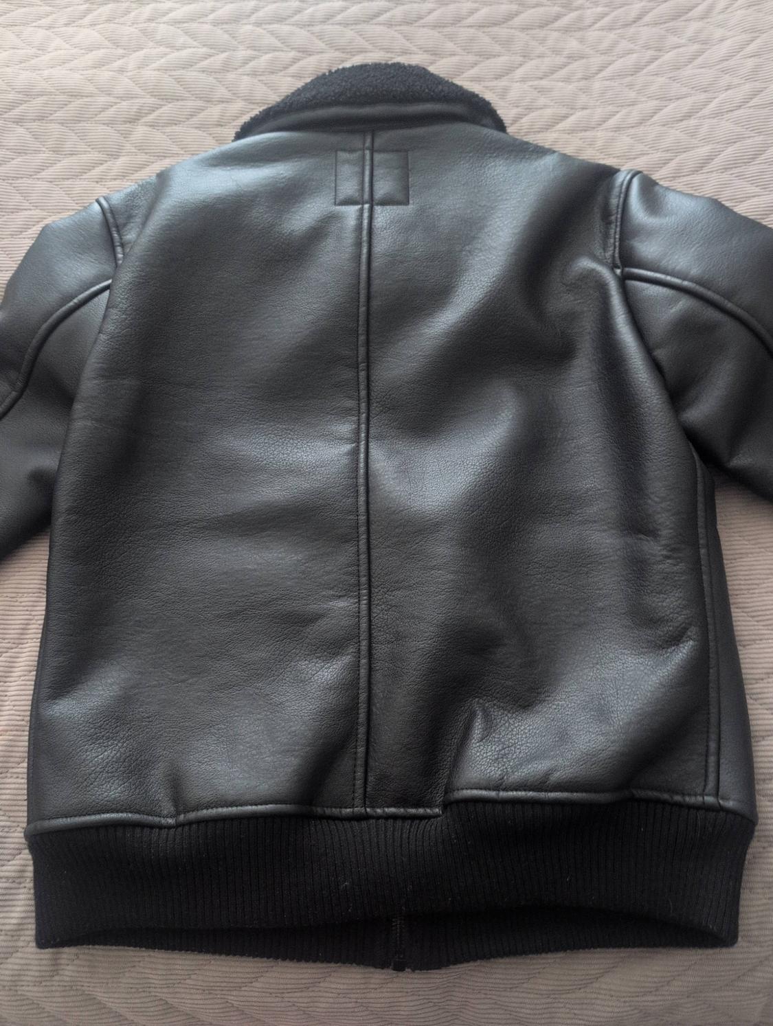 Black jacket viewed from the back with a textured collar, central seam and elasticated hem, on a beige surface with a zigzag pattern.